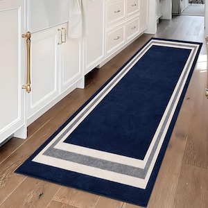 May include: A long rectangular runner rug featuring a deep navy blue center panel framed by concentric borders in light gray and off-white. The rug has a flat, uniform texture and is positioned lengthwise on a wooden floor in a kitchen setting.