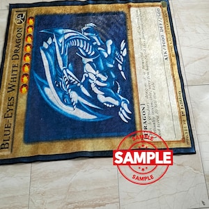 Może przedstawiać: Prostokątny dywan z Białym Smokiem Błękitnookim z gry karcianej Yu-Gi-Oh!. Dywan ma niebieskie tło z ilustracją białego smoka, złotą obw&oacute;dką i szczeg&oacute;łami karty.