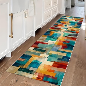 May include: A long rectangular runner rug featuring a vibrant abstract pattern of overlapping geometric blocks in shades of teal, deep blue, bright orange, yellow, and cream, laid out on a wooden floor in front of white kitchen cabinetry.