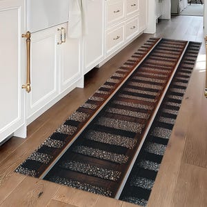 May include: A rectangular runner rug with a railroad track design. The rug features dark brown wooden railroad ties, gray gravel, and parallel silver rails. The rug is placed on a hardwood floor, with white cabinets and gold hardware in the background.