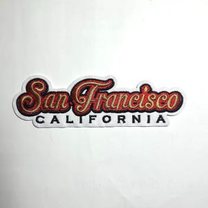 May include: Embroidered patch with the words "San Francisco CALIFORNIA" in a stylized font. The text is in red and gold, outlined in black, on a white background. The patch has a curved shape.
