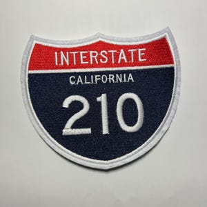 May include: Embroidered patch in the shape of a California Interstate highway sign. The patch features a red top section with the word "INTERSTATE" in white, a navy blue centre with "CALIFORNIA" and "210" in white, and a white border.