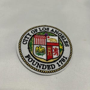 May include: Circular embroidered patch featuring the City of Los Angeles seal. The patch has a white background with a gold beaded border. The seal includes a shield with various emblems and the text "CITY OF LOS ANGELES FOUNDED 1781."