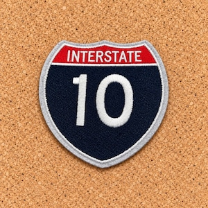 May include: A shield-shaped embroidered patch featuring the text INTERSTATE in white on a red background at the top, with the large white number 10 centered on a dark blue background below, all outlined with a light gray border.