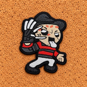Può includere: Una toppa ricamata raffigurante un personaggio dei cartoni animati che indossa un fedora nero, un maglione a righe rosse e nere e pantaloni bianchi. Il personaggio ha un viso color marrone chiaro con segni arancioni, un guanto nero con lame bianche e la lingua di fuori.