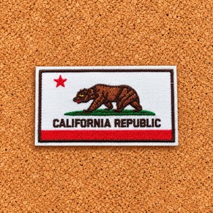 May include: A rectangular embroidered patch featuring the California state flag design. It displays a brown bear walking on a green patch of grass, a red star in the upper left corner, and the text CALIFORNIA REPUBLIC in brown block letters.