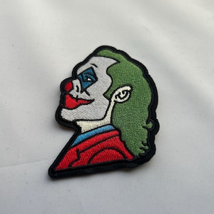 Pode incluir: Patch bordado com o perfil do rosto de um palhaço. O patch tem cabelo verde, maquiagem branca, nariz e boca vermelhos e maquiagem azul para os olhos. O palhaço usa um casaco vermelho com gola azul. O patch é contornado em preto.