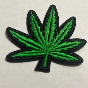 Könnte beinhalten: Gestickter Aufnäher in Form eines Cannabisblattes. Das Blatt ist leuchtend grün mit detaillierten Nähten, umrandet von Schwarz. Geeignet für Kleidung oder Accessoires.