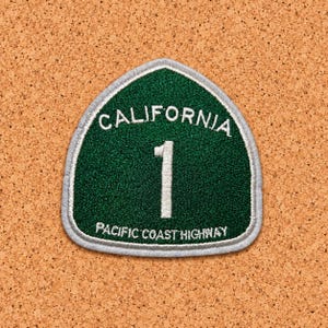 Puede incluir: Un parche bordado en forma de escudo con fondo verde oscuro y borde blanco. El texto CALIFORNIA aparece arqueado en la parte superior, un gran número 1 blanco en el centro y PACIFIC COAST HIGHWAY escrito en la parte inferior en blanco.