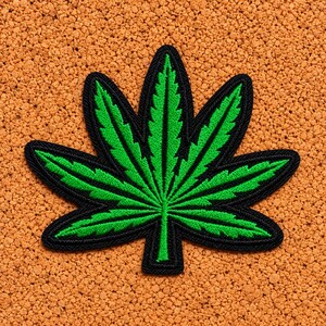 Puede incluir: Un parche bordado con forma de hoja de siete puntas que presenta un centro verde vibrante con costuras detalladas y un borde negro grueso. El diseño es simétrico y muestra los bordes dentados característicos de una forma de hoja.