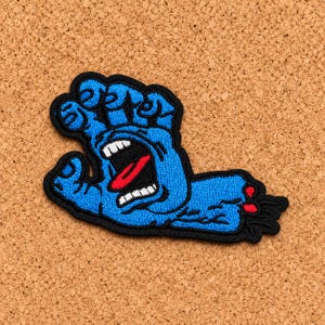 May include: An embroidered patch featuring a blue hand with a black outline. The hand has a wide-open mouth in the palm area, revealing white teeth and a red tongue. The wrist area is severed with red accents, and the entire design is bordered by black.