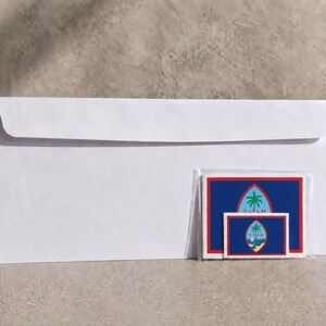 May include: A white business-sized envelope positioned behind a clear plastic sleeve containing two rectangular stickers featuring the blue, red, and white flag of Guam with a central emblem depicting a palm tree and a boat on the water.