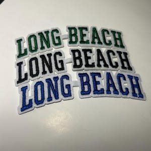 May include: Close-up of several embroidered patches with the text "LONG BEACH" in a collegiate font. The patches are in green, black, and blue, each with a white border. The patches are on a white surface.