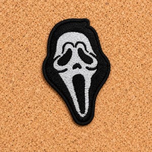 Op de afbeelding: Een geborduurde patch met het iconische witte spookmaskerontwerp met zwarte oogkassen, een kleine neus en een langwerpige open mond, allemaal omlijnd met zwart garen op een zwarte achtergrondvorm.
