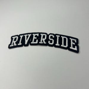 May include: A black and white embroidered patch with the word "RIVERSIDE" in white block letters. The letters are arched and outlined in black, with a white border. The patch is on a white surface.