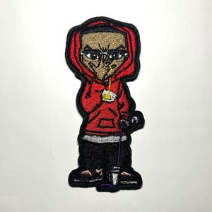 May include: Embroidered patch featuring a cartoon figure in a red hoodie and black pants. The figure has a stern expression and is holding a microphone. The patch has a black border and is likely for clothing or accessories.