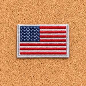 May include: A rectangular embroidered patch featuring the flag of the United States of America. The design includes a blue canton with white stars on the left and alternating horizontal red and white stripes on the right, finished with a light gray border.