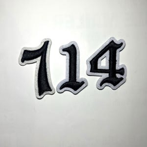 Könnte beinhalten: Drei schwarz-weiße Filz-Zahlenaufnäher. Die Zahlen sind in einem gotischen Schriftstil gehalten, mit schwarzer Mitte und weißem Rand. Die Zahlen sind 1, 1 und 4.