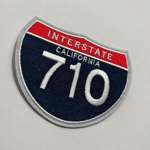 May include: Embroidered patch in the shape of a California Interstate highway sign. The sign is navy blue with white text reading "INTERSTATE CALIFORNIA 710". The top border is red, and the edge is grey.