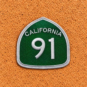 May include: A shield-shaped embroidered patch featuring a dark green background with a white border. The text CALIFORNIA is stitched in white at the top, and the number 91 is prominently displayed in large white digits in the centre.