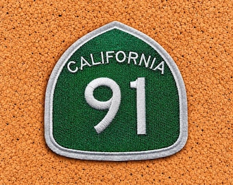 May include: A shield-shaped embroidered patch featuring a dark green background with a white border. The text CALIFORNIA is stitched in white at the top, and the number 91 is prominently displayed in large white digits in the center.