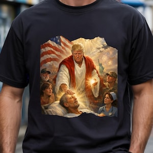 May include: Black t-shirt featuring a religious-themed graphic. The design depicts a figure in a red robe with a glowing hand, touching a person's forehead. An American flag and the Statue of Liberty are in the background.