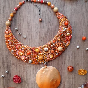 Könnte beinhalten: Eine halbmondförmige Perlenkette mit einem großen, runden, orangefarbenen Muschelanhänger. Die Kette besteht aus orangefarbenen, braunen und silbernen Perlen. Die Kette liegt auf einer Holzoberfläche mit kleinen orangefarbenen und silbernen Perlen, die um sie herum verstreut sind.