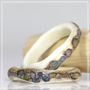 Puede incluir: Dos anillos con una base lisa de color blanco roto que presenta una banda texturizada en tonos marrones, incrustada con pequeñas piezas pulidas similares a piedras en tonos verdes, azules y morados, creando un aspecto orgánico y moteado.