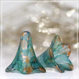 May include: Two turquoise and gold-colored glass flower-shaped beads. The beads have a flared, petal-like design with a metallic gold finish. The beads are set against a blurred, light-colored background.