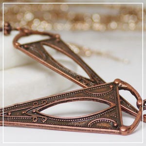 May include: Close-up of two ornate, copper-colored metal hair accessories. Each piece has a triangular shape with a central oval opening and detailed patterns. The background is blurred, suggesting a soft, neutral surface.