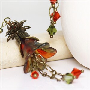 May include: A handcrafted floral pendant with a bronze finish. The pendant features a detailed flower with red and orange hues, accented by dangling red and green crystal beads. The design includes intricate chain details.