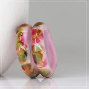 May include: Two smooth, rounded rings crafted from polished glass. The rings feature a swirling, marbled pattern of soft pink, muted green, and warm amber tones. The surface is glossy and reflective, showing light highlights across the curved bands.