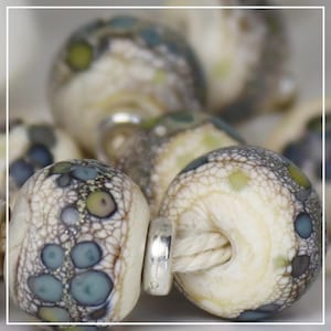 May include: Close-up of artisan beads with a cream-colored base, speckled with blue, green, and gray patterns. The beads have a textured, web-like surface and are strung on a natural-colored cord. A silver-colored metal bead is used to secure the cord.