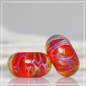 May include: Two cylindrical glass beads featuring a vibrant red base color with swirling patterns of white and purple throughout the surface. The beads have a smooth, glossy finish and a central hole, reflecting light on their rounded, polished edges.