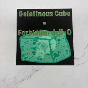 May include: A black square sticker with green text that reads "Gelatinous Cube = Forbidden Jell-O." The sticker features a green, cube-shaped illustration with a skull inside. The cube has green splatters.