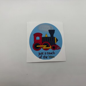 May include: A blue and white circular sticker with a cartoon red and black train engine. The text "just a touch of the 'tom" is below the train.