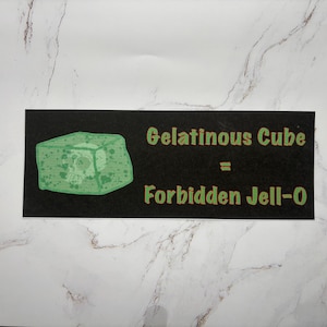Könnte beinhalten: Ein schwarzes rechteckiges Schild mit dem Text "Gelatinous Cube = Forbidden Jell-O" in grünen Buchstaben. Auf der linken Seite des Schildes befindet sich eine grüne, würfelförmige Illustration. Das Schild befindet sich auf einer weiß-grau marmorierten Oberfläche.