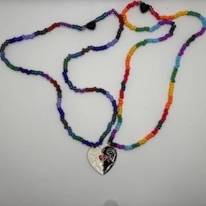 May include: A two-piece necklace set featuring small, multi-coloured beads. One strand is primarily blue, purple, and green, while the other is a rainbow gradient. Each strand connects to a half-heart pendant that forms a complete heart when joined.