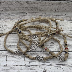 May include: A collection of hemp cord bracelets, each featuring a silver heart charm and colourful beads. The bracelets are a natural tan colour, with a rustic, handmade look. The beads are red, blue, and yellow.
