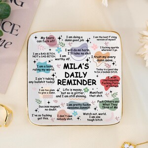 Könnte beinhalten: Ein weißer quadratischer täglicher Reminder mit dem Text "MILA'S DAILY REMINDER" und verschiedenen Affirmationen. Der Reminder ist von Goldschmuck und Blumen umgeben. Der Text enthält Sätze wie "Ich bin ein Boss, der meine Welt regiert."