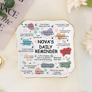 Könnte beinhalten: Eine weiße Keramik-Tageserinnerungsschale mit Goldrand, die verschiedene positive Affirmationen und den Text "Nova's Daily Reminder" enthält. Die Schale ist mit Blumen- und Sternmotiven verziert.