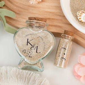 Pode incluir: Duas garrafas de vidro cheias de areia, uma em forma de coração e outra cilíndrica, com rolhas. A garrafa em forma de coração tem os nomes "Linda & Kelly" e "Hawaii 06.12.2025" impressos, juntamente com as iniciais "K & L" e um desenho floral. A garrafa cilíndrica tem a data "10.14.25" impressa.