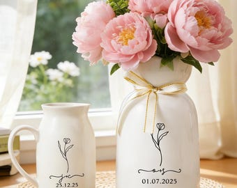 May include: A white ceramic mug and a matching white ceramic vase filled with pink peony flowers. Both items feature a black line drawing of a single flower, the initials "jc" and "as", and the dates "25.12.25" and "01.07.2025" respectively.
