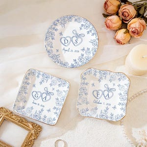 Puede incluir: Tres platos de cerámica blanca con motivos florales azules y bordes dorados. Cada plato presenta iniciales dentro de corazones enlazados y una fecha. Se muestra un plato redondo, un plato cuadrado y un plato festoneado.
