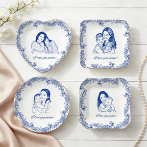 May include: Four white ceramic plates with blue floral designs and gold rims. Each plate features a blue sketch of a mother and child, with the text "I love you mom!". The plates are in heart, square, round, and square shapes.