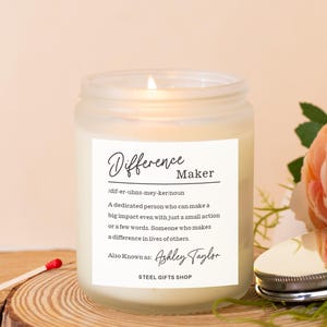 Op de afbeelding: Een brandende, heldere glazen kaarsenpot met een wit etiket met de tekst "Difference Maker". Het etiket bevat een definitie en de naam "Ashley Taylor". Een lucifer en een bloem staan naast de kaars op een houten oppervlak.