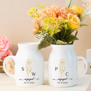 May include: Two white ceramic mugs with handles, featuring a gold floral design and the initials "SW" and "PC" with the text "engaged 03.16.2025". The mugs are filled with a bouquet of yellow and peach flowers.