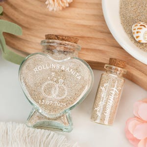 May include: Two glass bottles filled with sand, one heart-shaped and one cylindrical, with cork stoppers. The heart-shaped bottle has the names "Rollins & Anita" and the date "12.07.2025" printed on it, along with wedding rings. The cylindrical bottle has the name "Williams" printed on it.