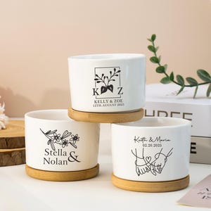 May include: Three white ceramic plant pots with wooden saucers. Each pot has a black graphic and text: "KELLY & ZOE 12TH AUGUST 2025", "Stella & Nolan", and "Keith & Maria 02.20.2025". The pots are cylindrical and sit on round wooden saucers.