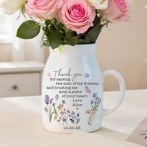 May include: A white ceramic pitcher with a handle, filled with pink and white roses. The pitcher has a floral design and the text "Thank you for raising the man of my dreams... Love Alice 12.20.25". A small vase of white flowers is to the right.